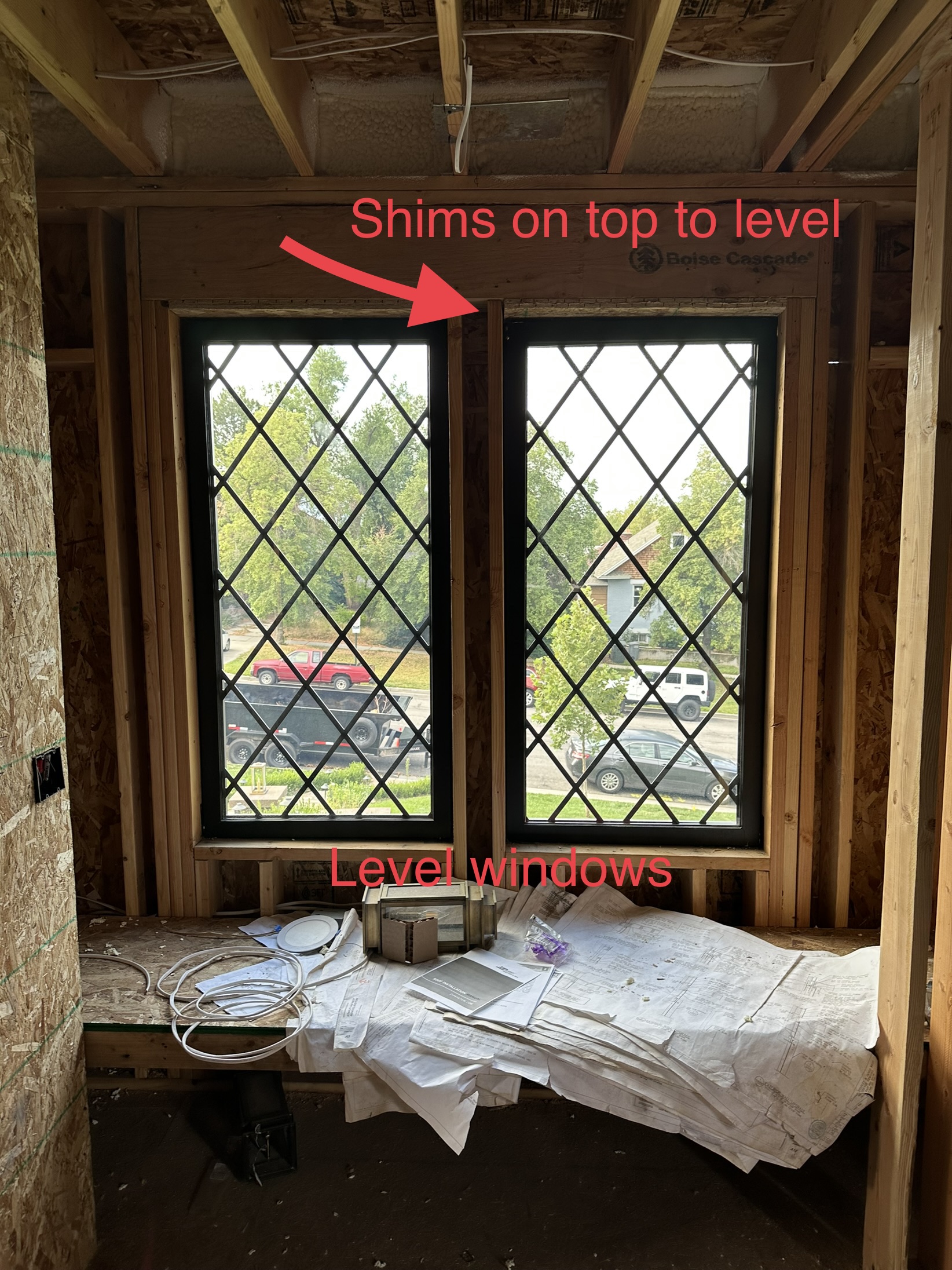 Annotated construction photo showing diamond-pane windows with red arrows pointing to shims on top to level — documenting precision work in an out-of-square structure