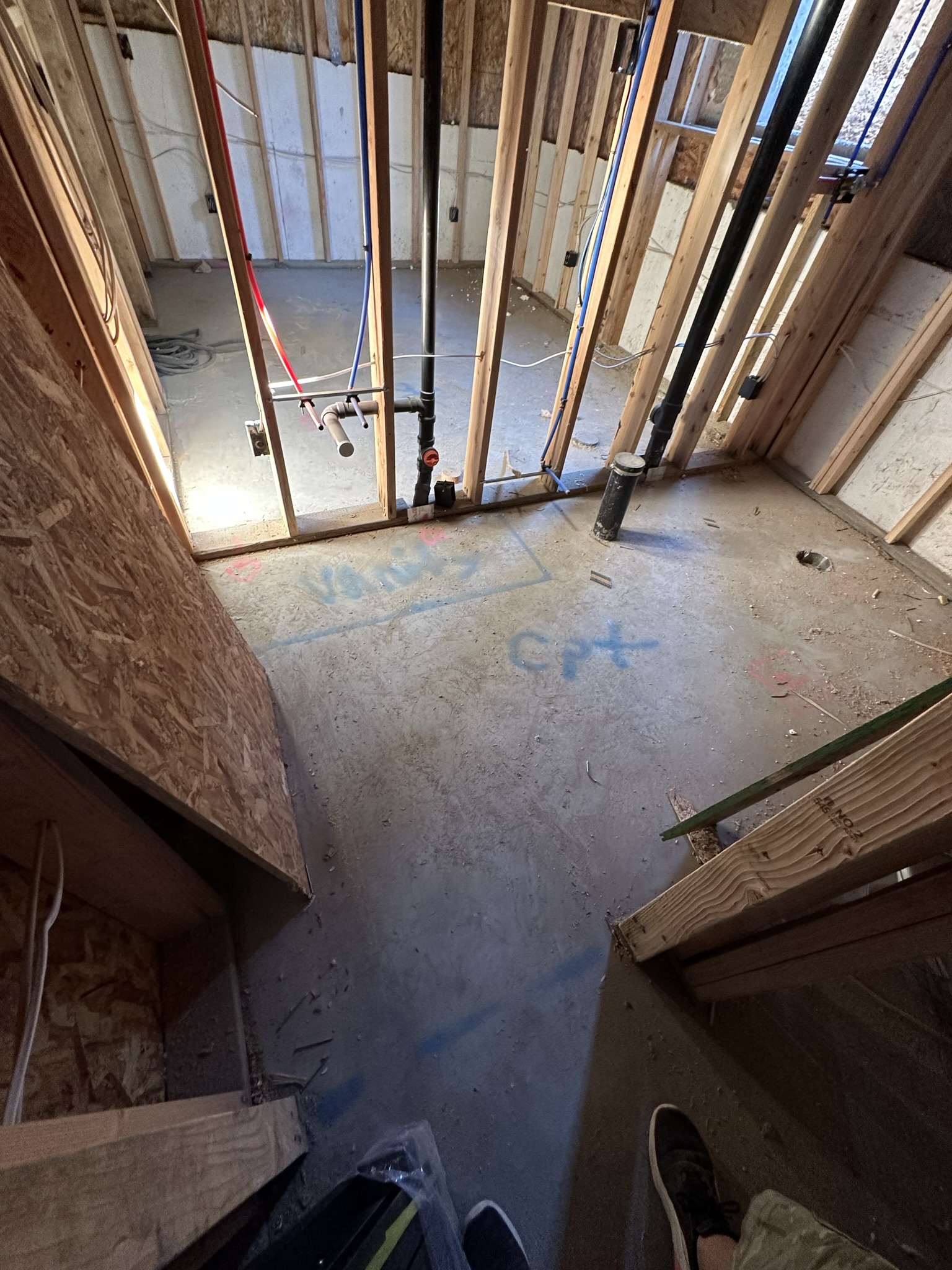 Looking down into basement showing PEX plumbing and ABS drain pipes — spray paint markings on concrete floor for layout coordination