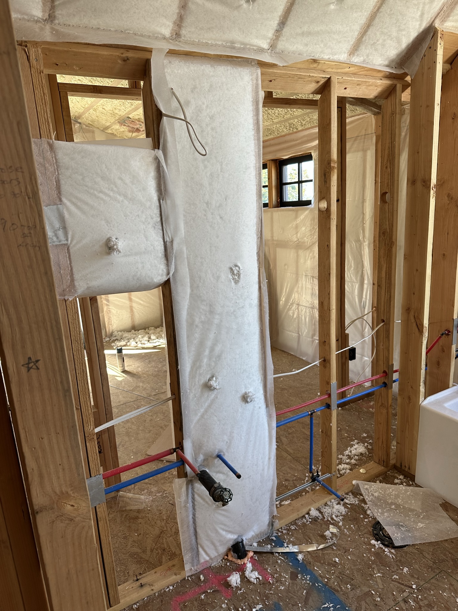 Fiberglass batt insulation being installed around PEX plumbing lines — vapor barrier visible, interior walls being insulated