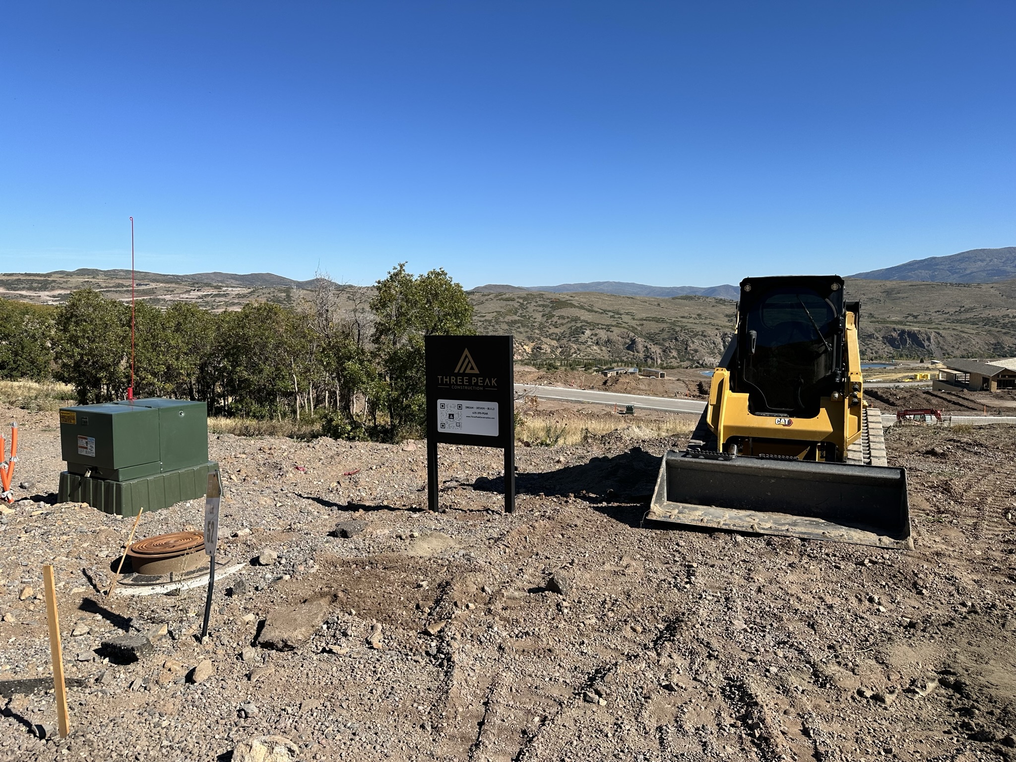 Three Peak Construction job site sign on the raw lot with CAT skid steer and Jordanelle Reservoir behind
