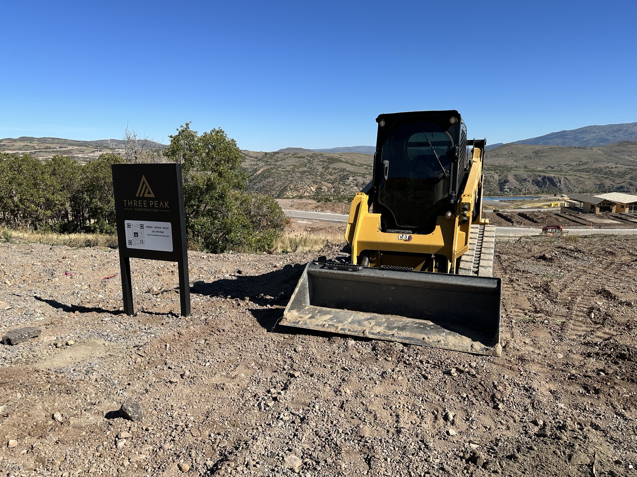 Close-up of Three Peak Construction sign — Dream, Design, Build — with equipment and reservoir visible
