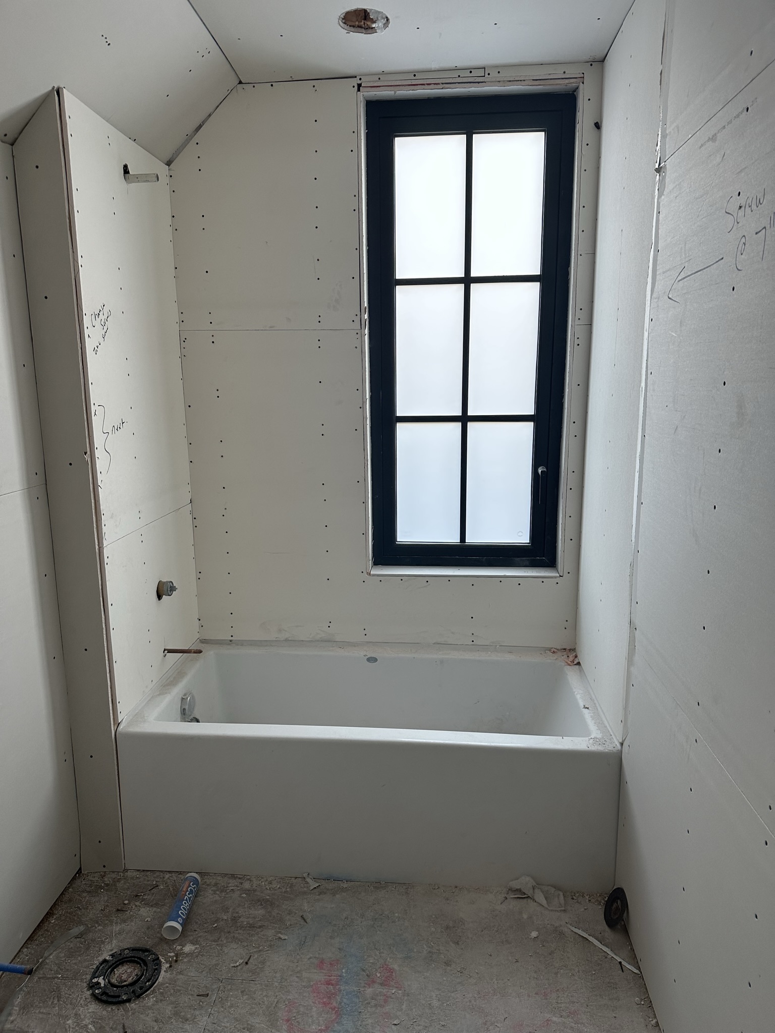 Bathroom taking shape — tub set in place, drywall hung, black-frame window installed, plumbing complete