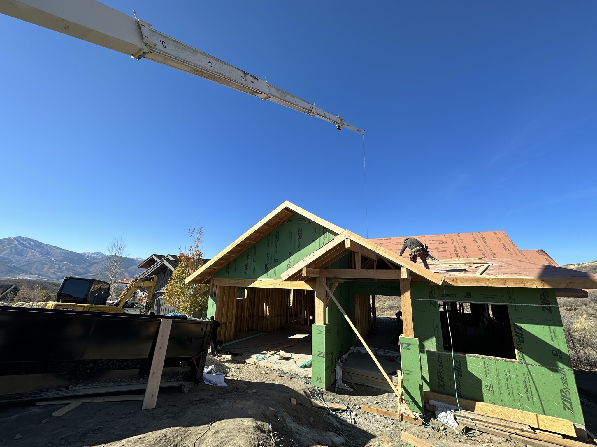 Wide shot of house framing with roof trusses, crane arm visible, and mountains in the background