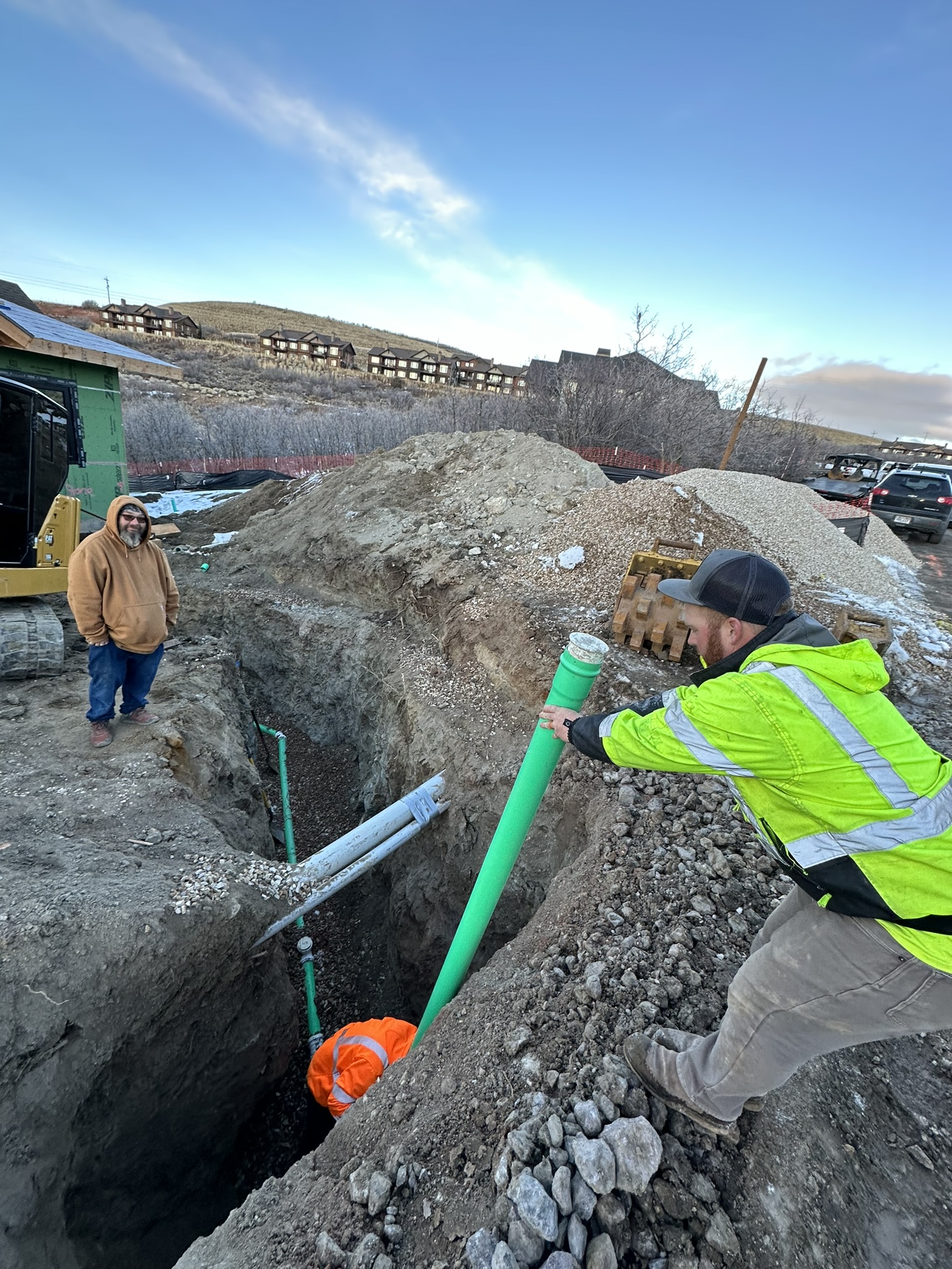 Workers installing sewer pipe in excavated trench with Hideout community visible in background