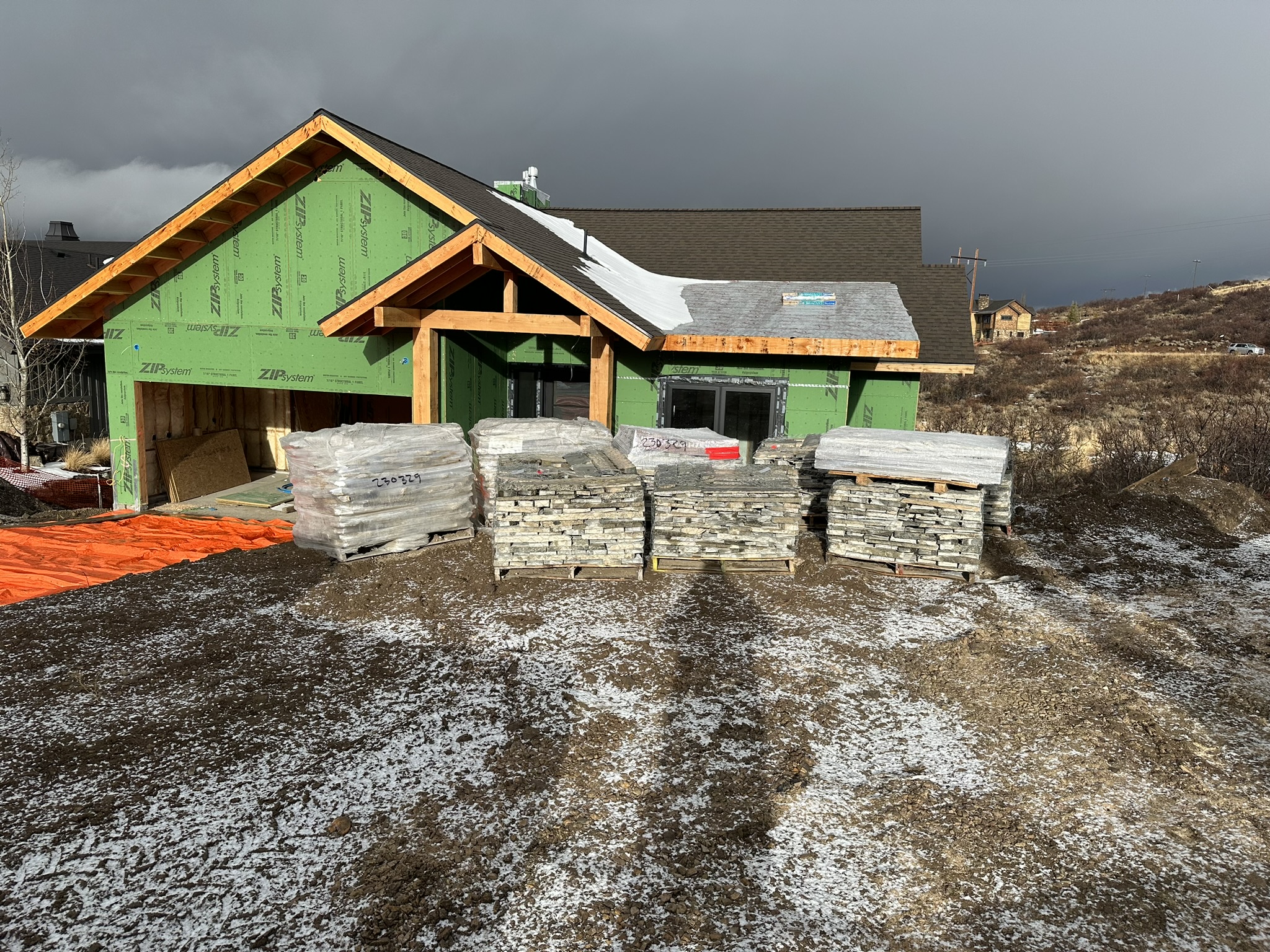 Front view of the home during framing — ZIP system sheathing, timber entry frame, roof trusses, stone pallets staged in foreground