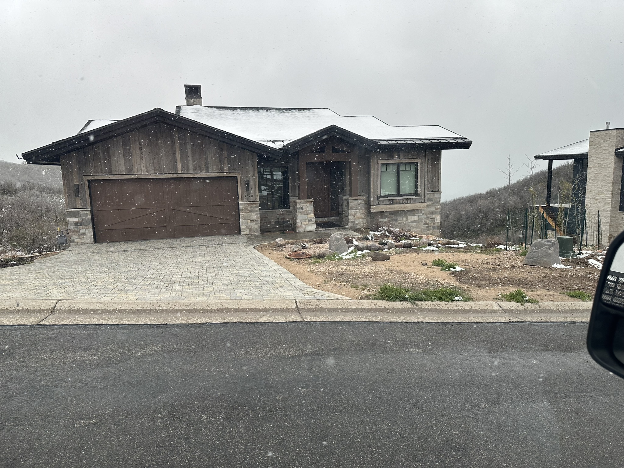 Near-complete exterior in snow — wood siding, stone columns, paver driveway, garage door installed