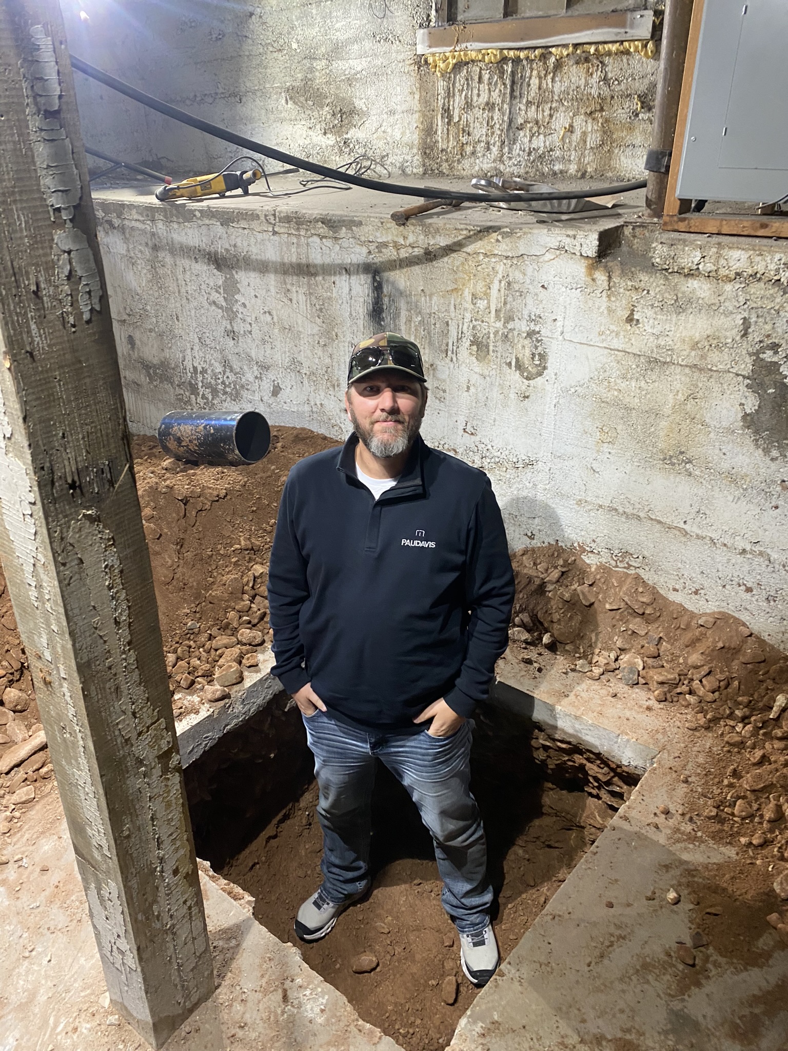 Nate Jensen standing in a deep footing excavation pit next to the foundation wall — demonstrating the depth of underpinning work required for structural repair