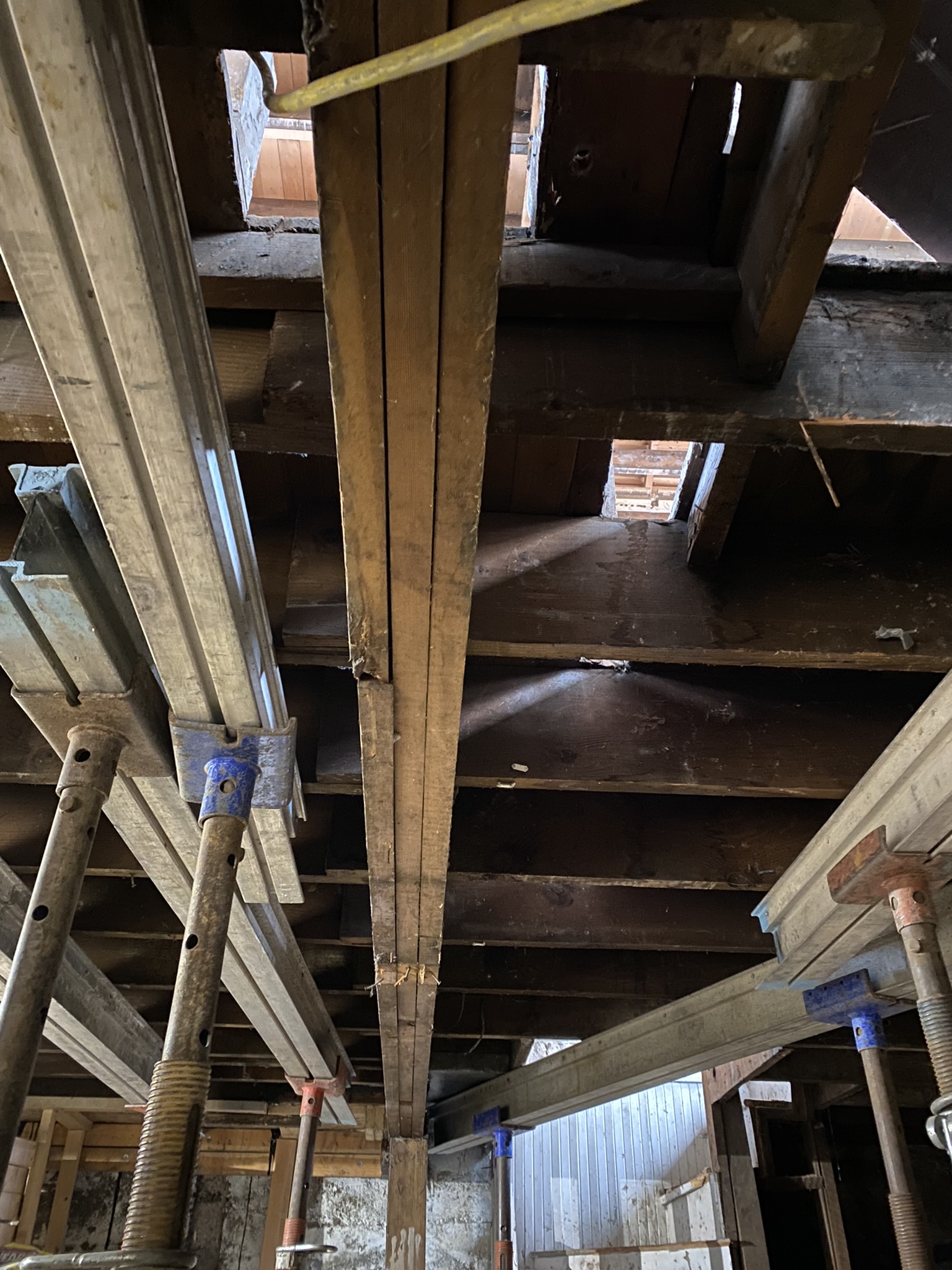 Looking up through floor joists at temporary steel shoring system — gaps in subfloor visible showing the extent of structural compromise