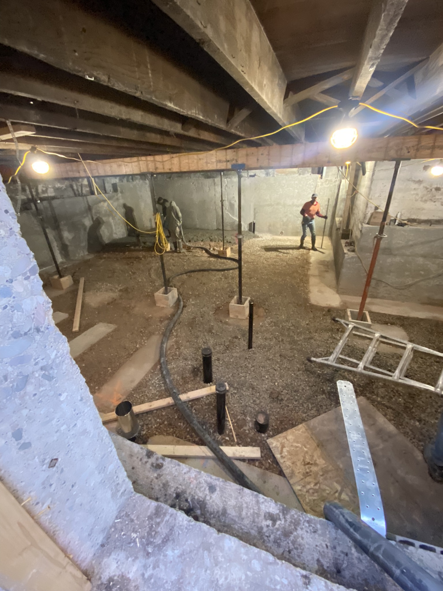 Wide view of crawlspace being rehabilitated — new concrete walls poured, steel posts on pad footings, worker in orange hi-vis