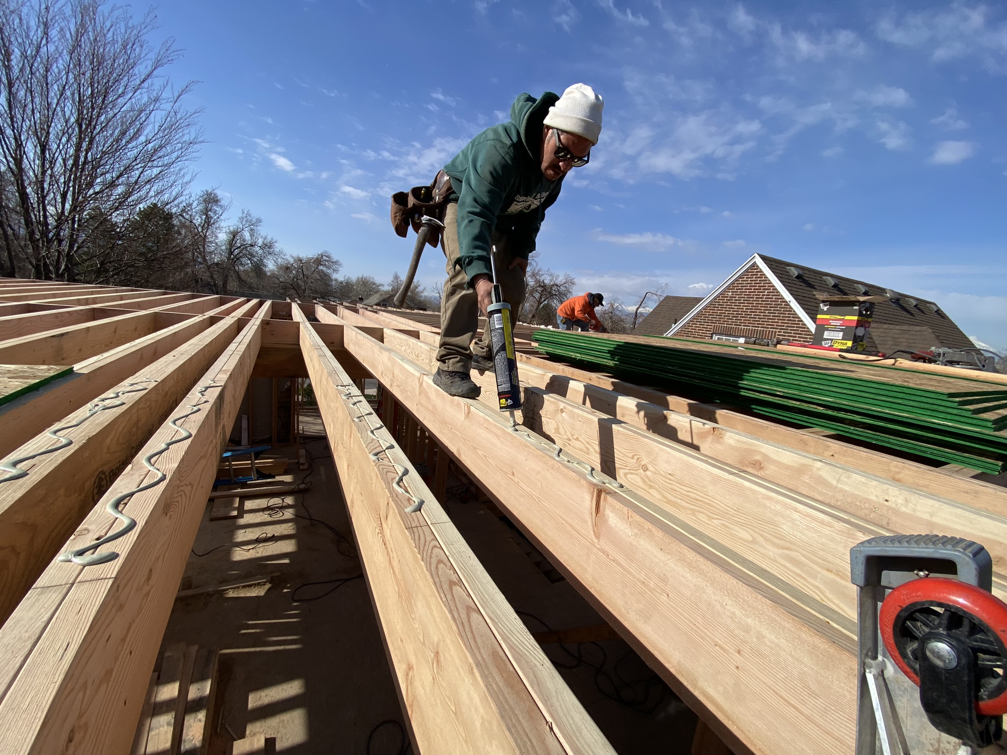 Worker with caulk gun sealing new floor joists — mountains visible in background, precision craftsmanship in every connection