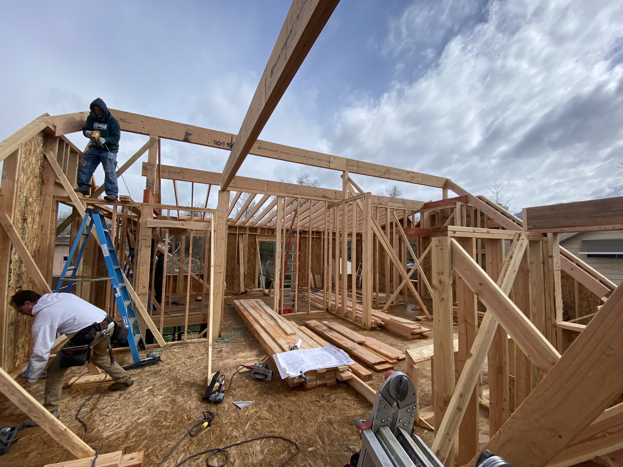 Wide angle of upper level framing — workers on ladders, ridge beam visible, blueprints on floor, dramatic clouds through open walls