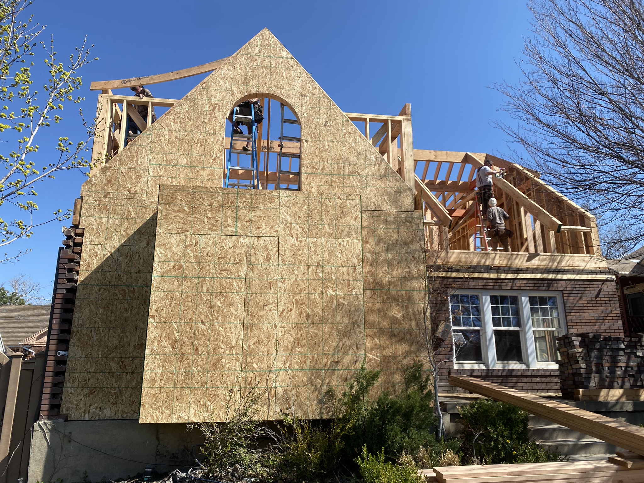 Full exterior view of new gable end wall with arched window opening — OSB sheathing complete, workers on ladders, massive scale of addition visible next to original brick