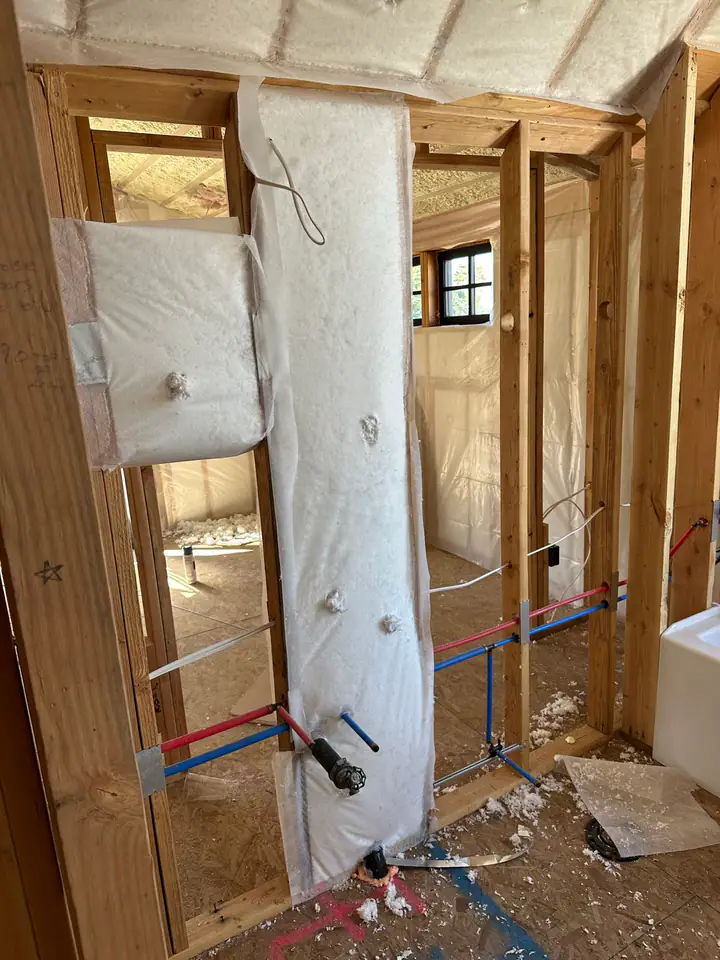 Fiberglass batt insulation being installed around PEX plumbing lines — vapor barrier visible, interior walls being insulated