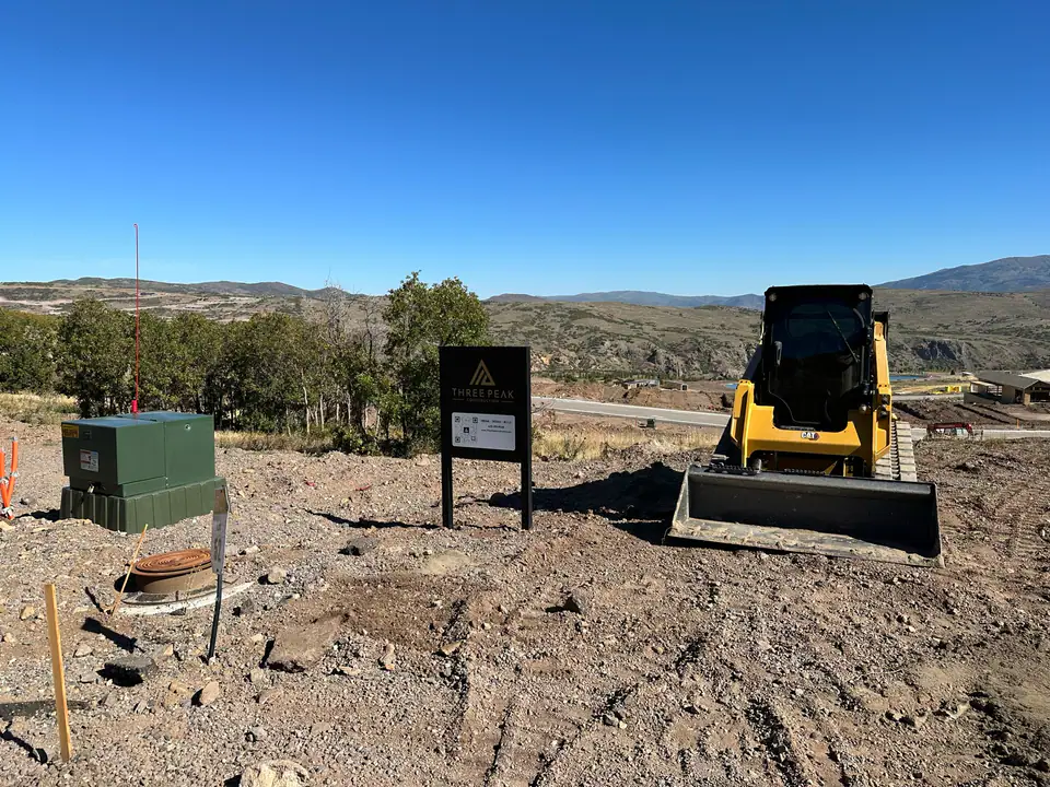 Three Peak Construction job site sign on the raw lot with CAT skid steer and Jordanelle Reservoir behind