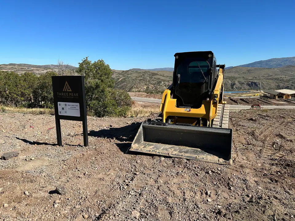 Close-up of Three Peak Construction sign — Dream, Design, Build — with equipment and reservoir visible