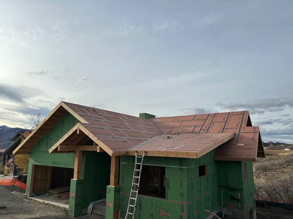 Full exterior view of the home during framing — ZIP System green sheathing on walls, ZIP-R roof panels, timber gable entry, ladder against the side, fall colors on the hillside