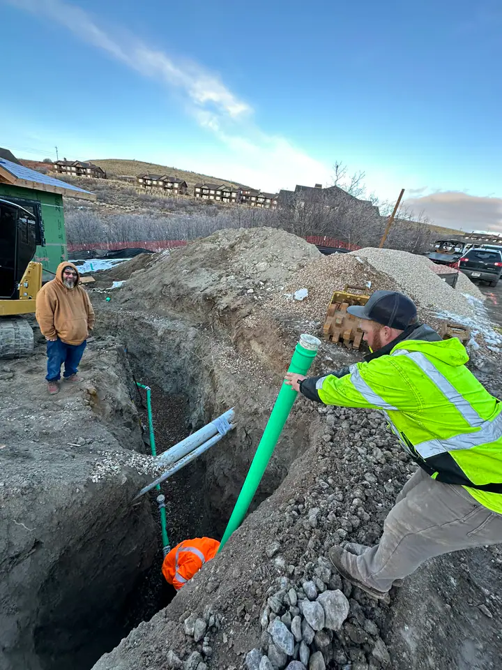 Workers installing sewer pipe in excavated trench with Hideout community visible in background