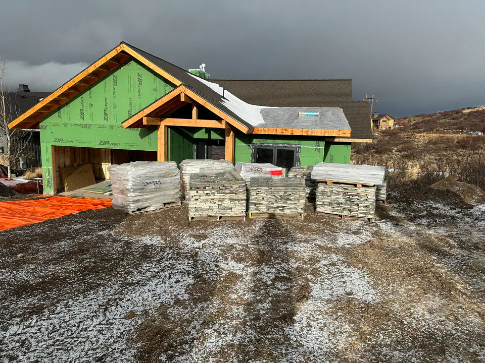 Front view of the home during framing — ZIP system sheathing, timber entry frame, roof trusses, stone pallets staged in foreground