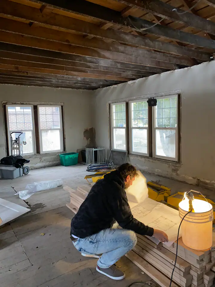 Nate Jensen reviewing structural blueprints inside a gutted older home — exposed ceiling joists and natural light through original windows