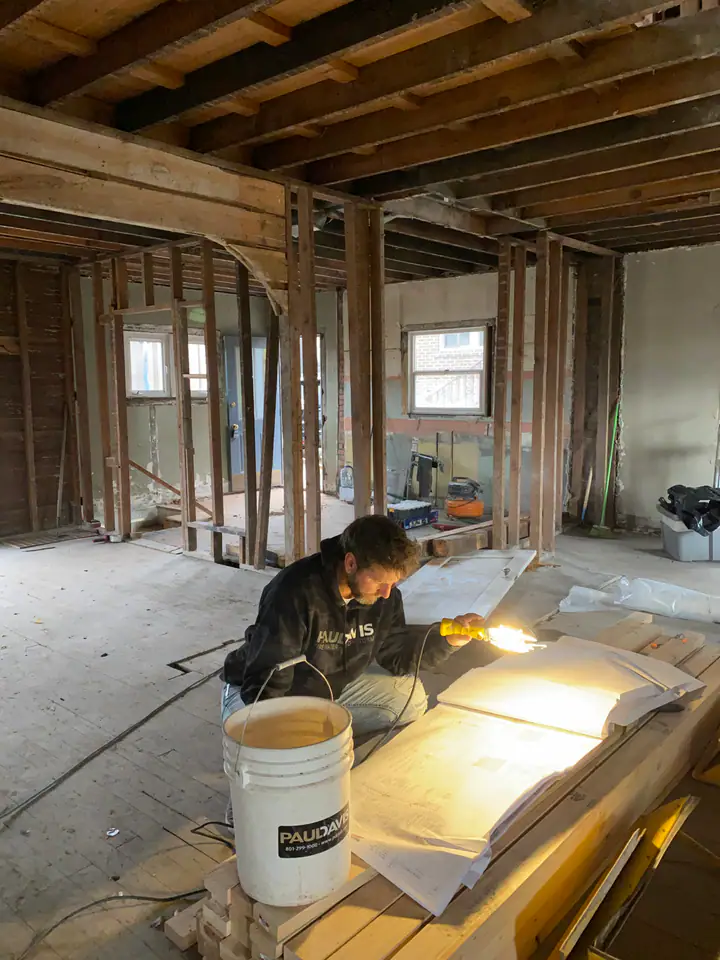Nate reviewing blueprints by work light in a fully framed room during structural rehabilitation