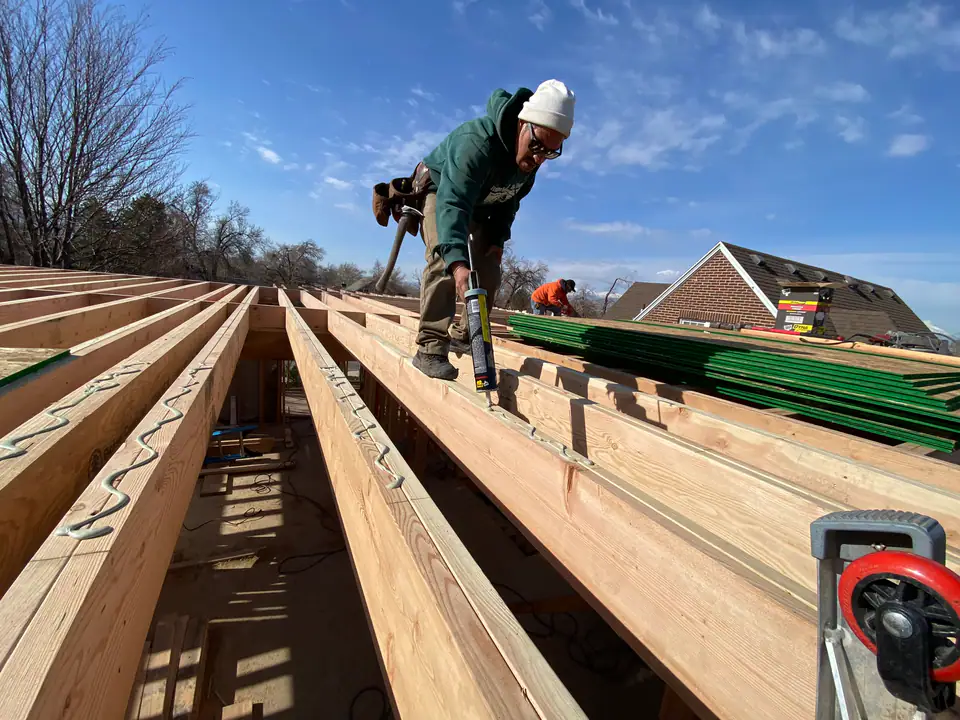 Worker with caulk gun sealing new floor joists — mountains visible in background, precision craftsmanship in every connection